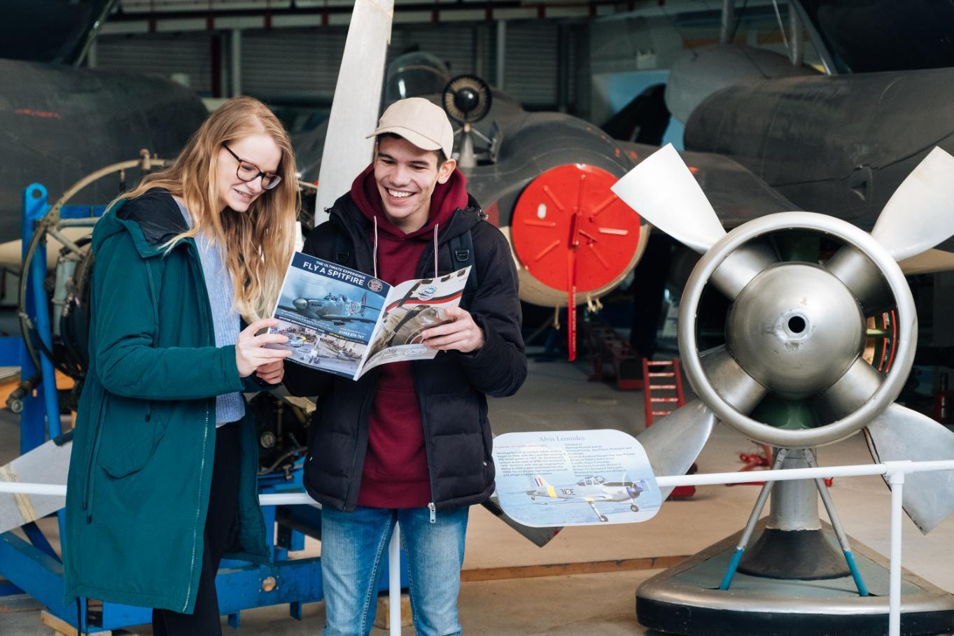 Two people looking at a leaflet and smiling stood in front old plane at Solent Sky musuem