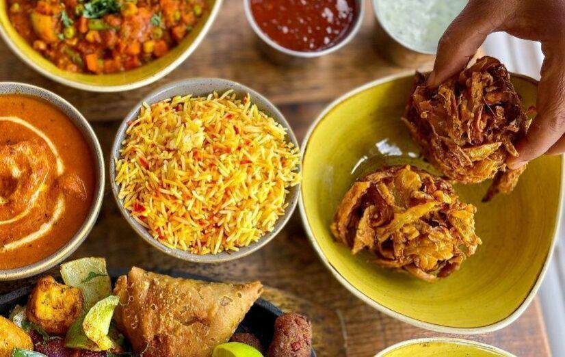 Hand grabbing beer battered onion bhaji from yellow dish next to spread of colourful Indian dishes that include rice pilau, chicken curry and samosa