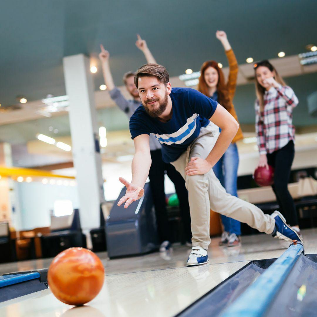 Man throwing orange bowling ball down bowling lane as friends cheer behind him